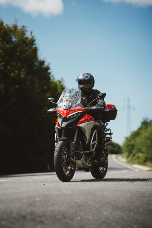 Motociclista com capacete numa moto de turismo vermelha, numa estrada com céu azul e árvores